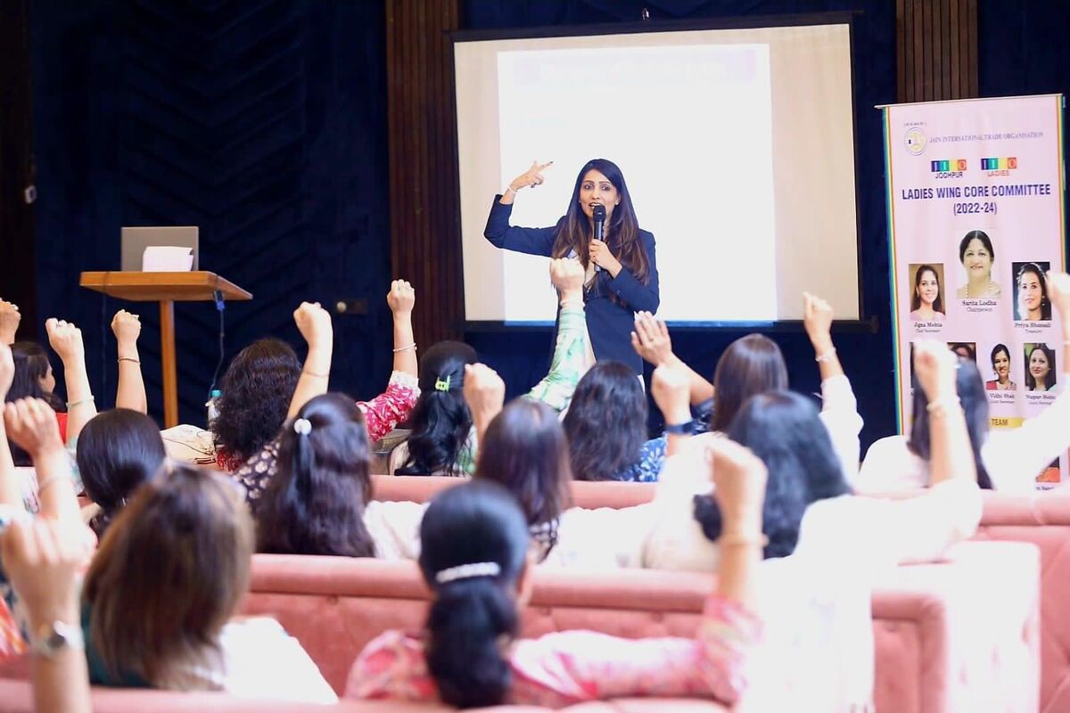 Prerna Maheshwari speaking on stage with a microphone at a JITO seminar, audience members with raised fists in the foreground