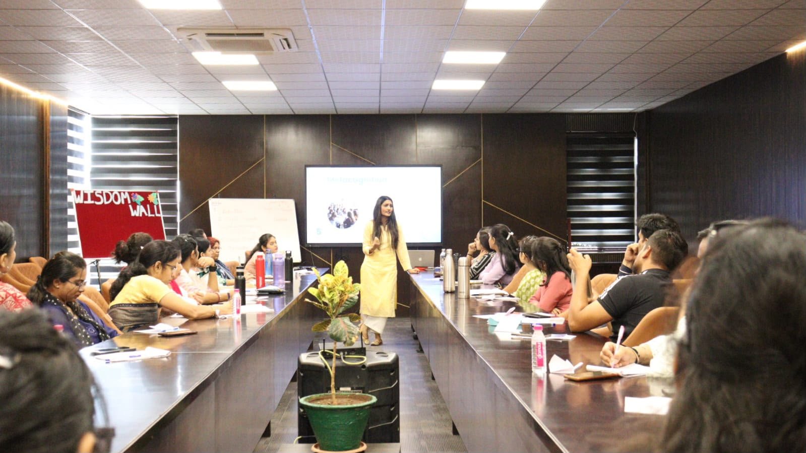 Prerna Maheshwari presenting in a corporate boardroom beside a "Wisdom Wall" display